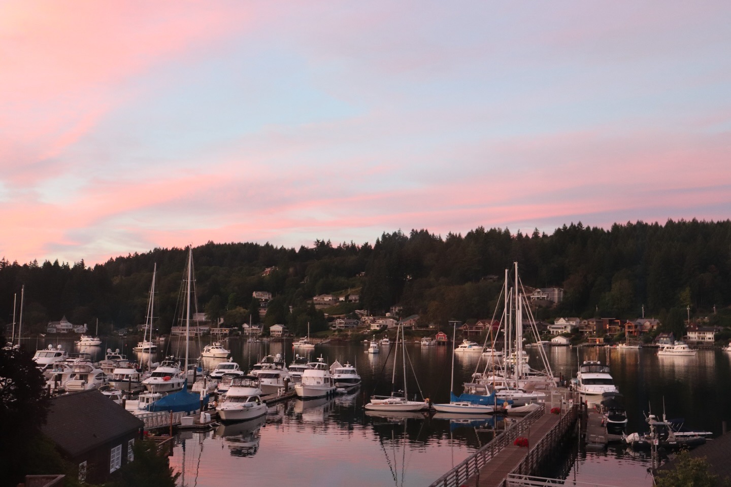 Gig Harbor marina at dusk under a soft pink sky, boats moored along the docks with forested hillsides beyond