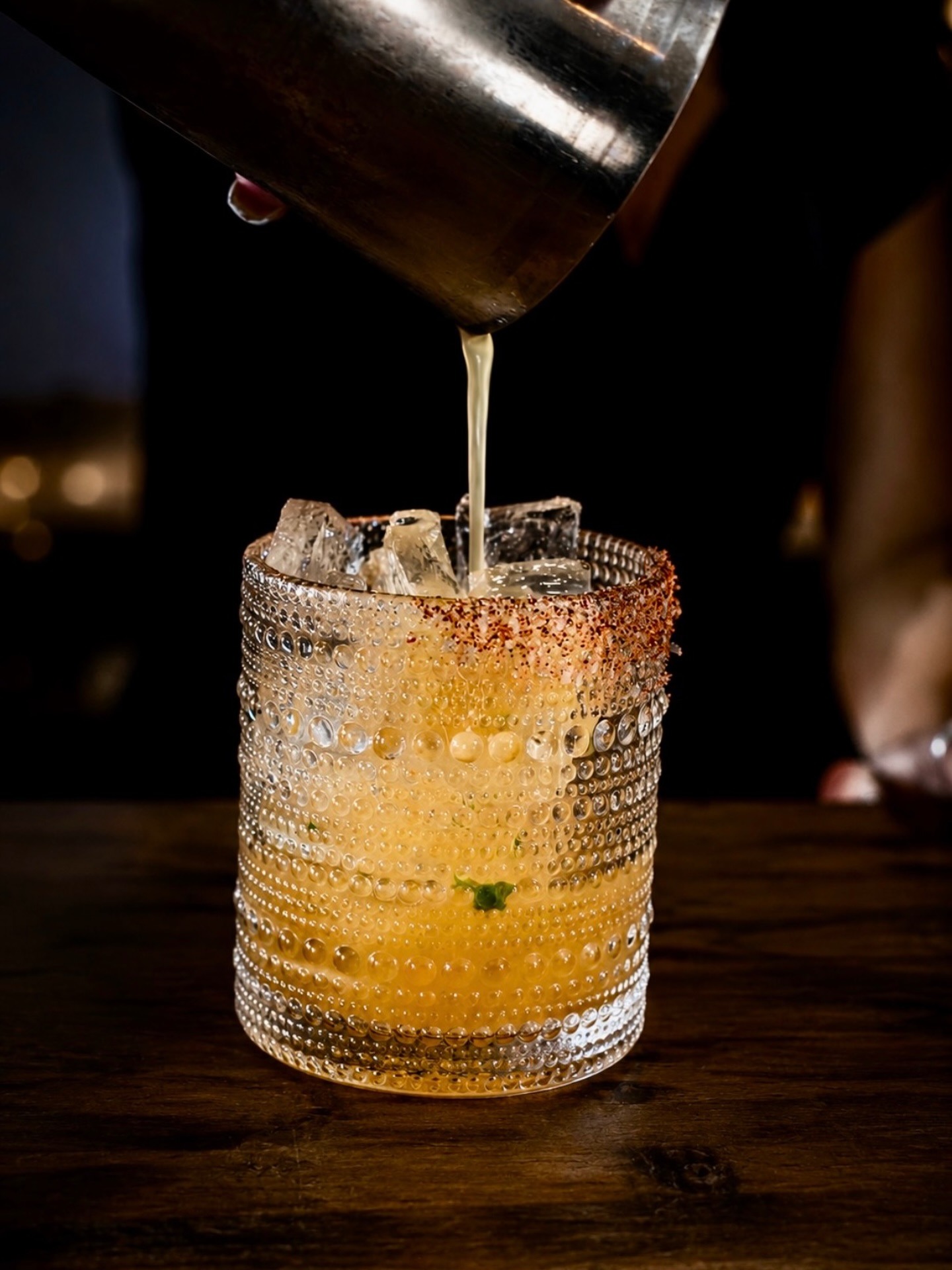 A bartender straining a freshly shaken cocktail into a chili-salt-rimmed hobnail glass over ice at the Brix 25° bar