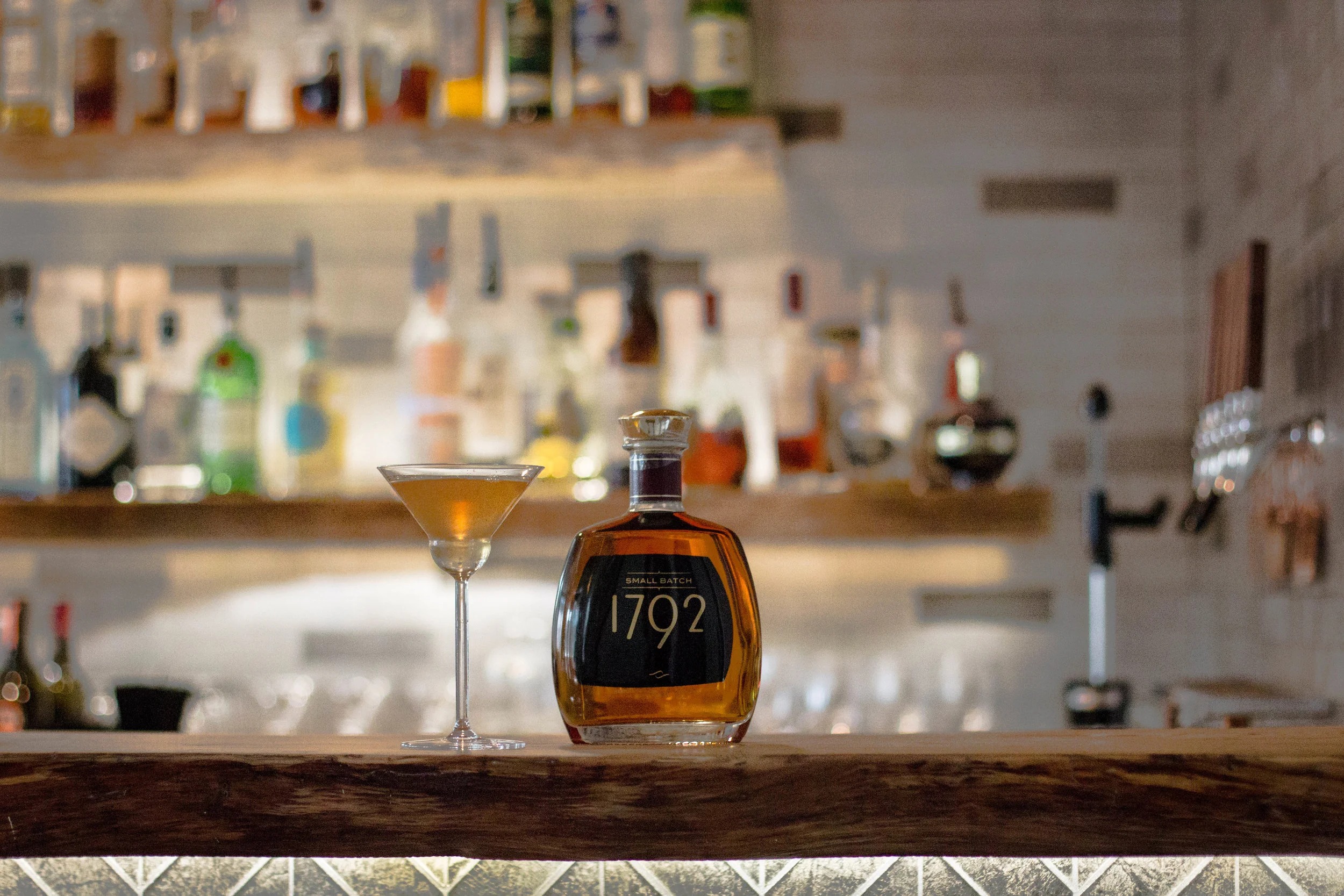 A small-batch bourbon bottle and a stirred cocktail in a coupe on the live-edge wood bar at Brix 25°, backed by softly lit shelves of spirits