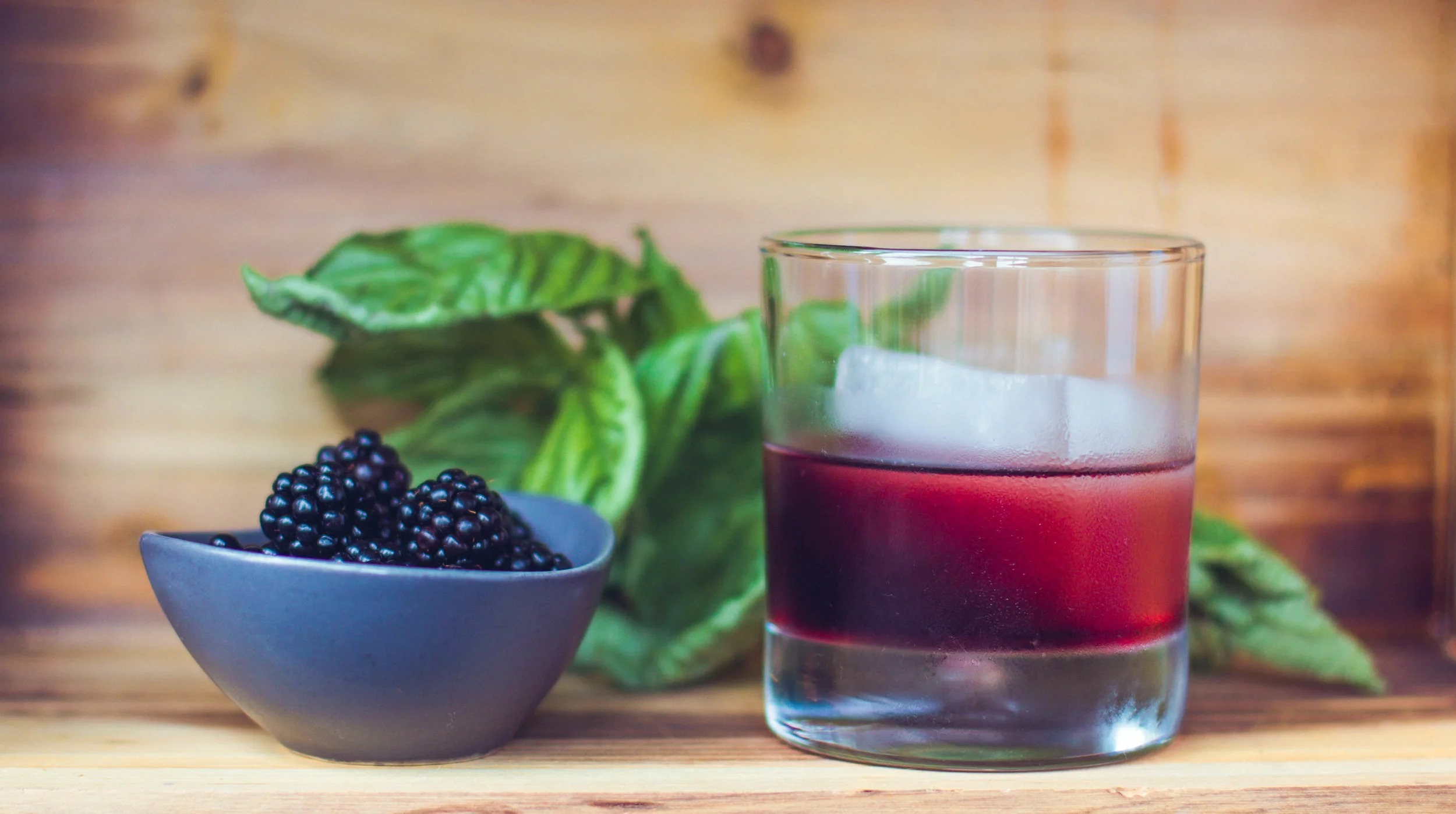 A deep-berry cocktail over a single large ice cube beside a bowl of fresh blackberries and basil leaves on a wood table at Brix 25°
