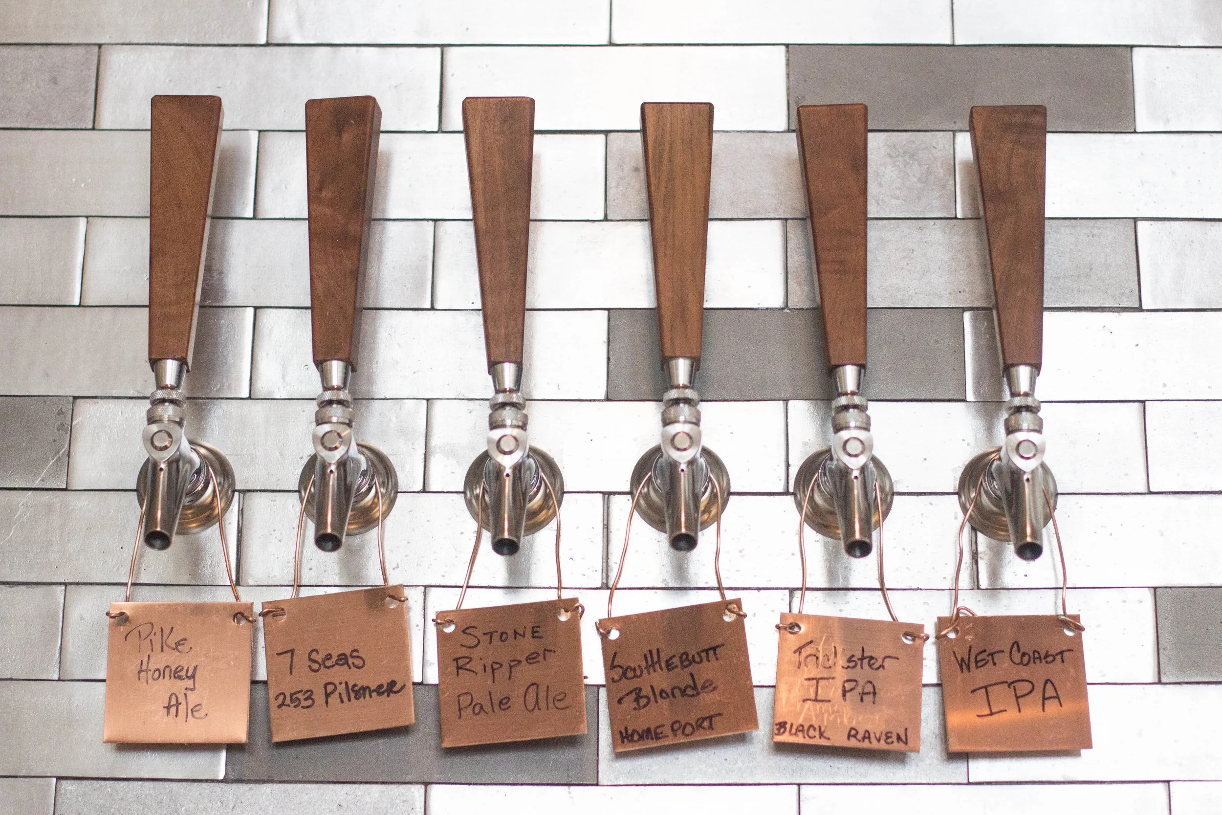Six wooden draft tap handles with hand-lettered copper tags for rotating Northwest beers at Brix 25°