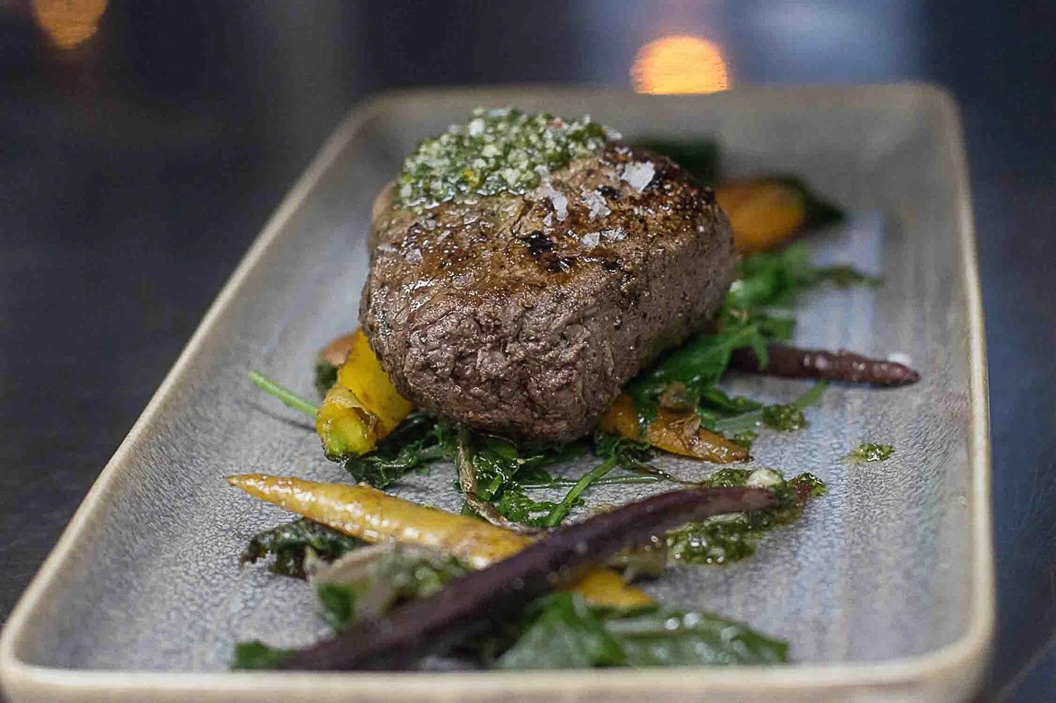 A seared Prime steak with chimichurri and heirloom carrots, plated over greens by candlelight at Brix 25°